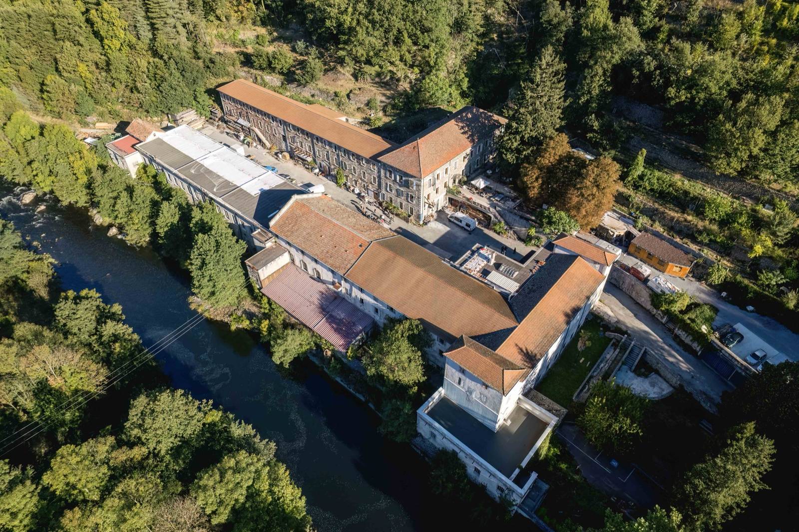 Des bâtiments anciens au bord d'une rivière, dans une vallée boisée, vus du ciel au drone
