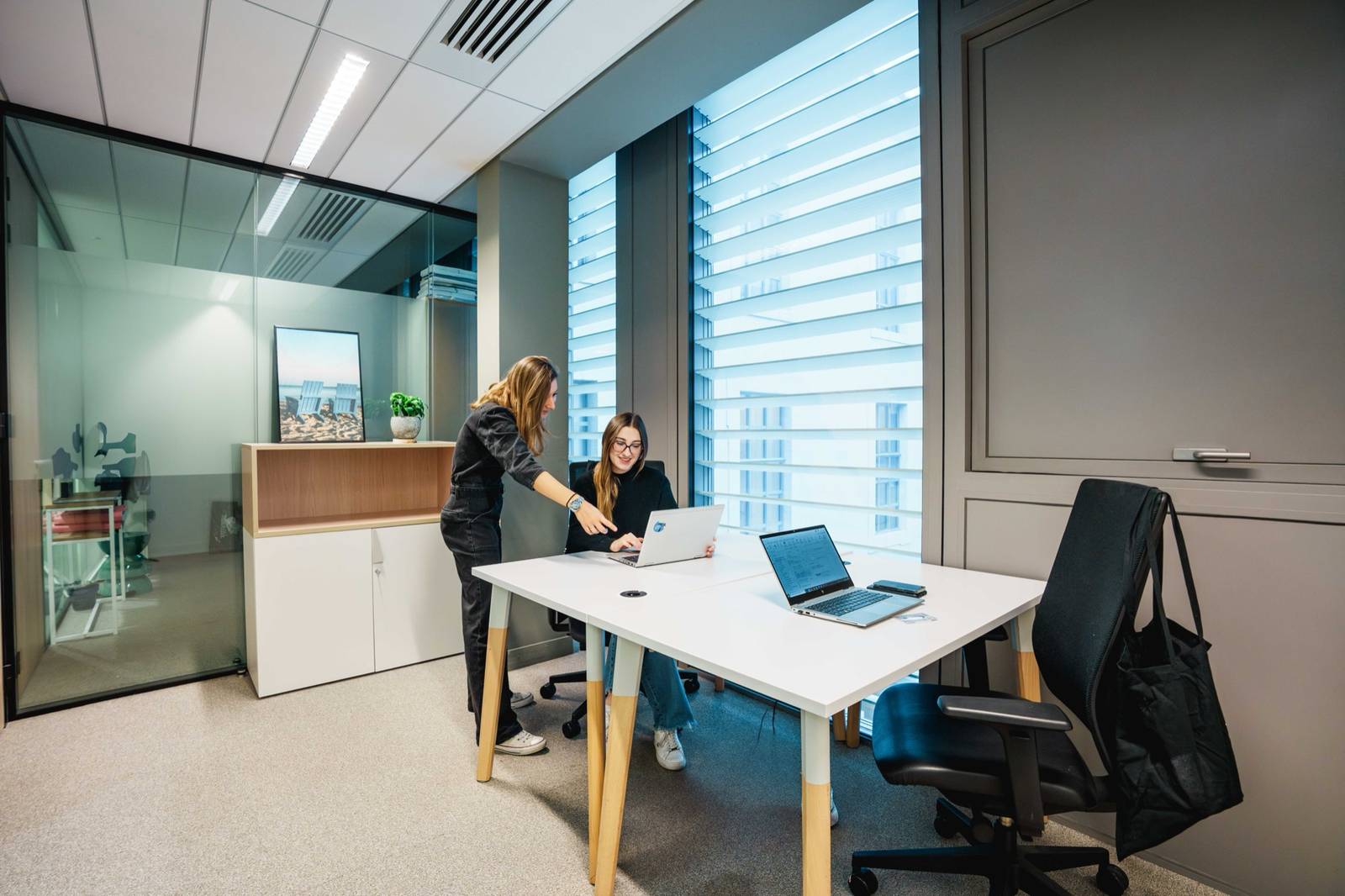 Deux collègues échangent devant un ordinateur dans un petit bureau vitré