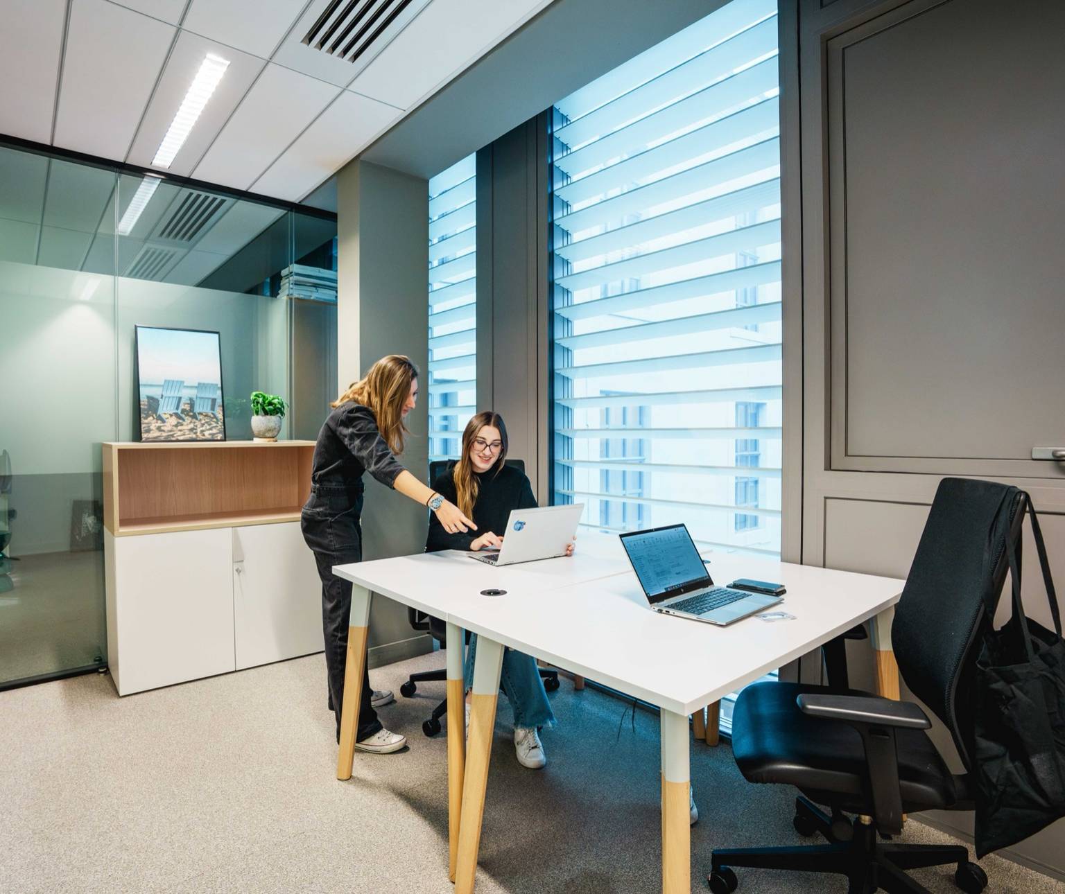 Deux collègues échangent devant un ordinateur dans un petit bureau vitré