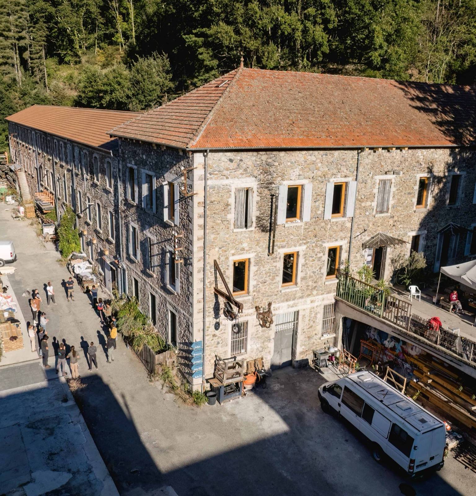 Un grand bâtiment en pierre et sa cour animée, dans une vallée boisée, vus en surplomb au drone
